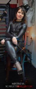 Photo of Mistress B Wilde sititng crossed legged on high timber chair. She is wearing leather kink body suit and black high heels. Mistress holds a straight crop in her hands.