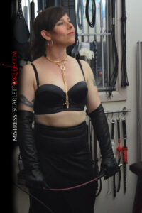 Photo of Mistress B Wilde in leather skirt and gloves against a kink dungeon wall, with lots of whips and BDSM toys