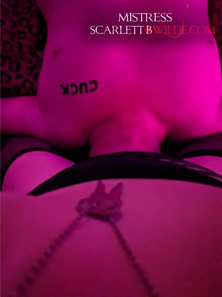 Photo in POV style as slave lies under Mistress on a bed- ‘cuck’ tattooed on his chest. He eats Mistress's pussy like a good boy, as a key to his cage hangs from her necklace