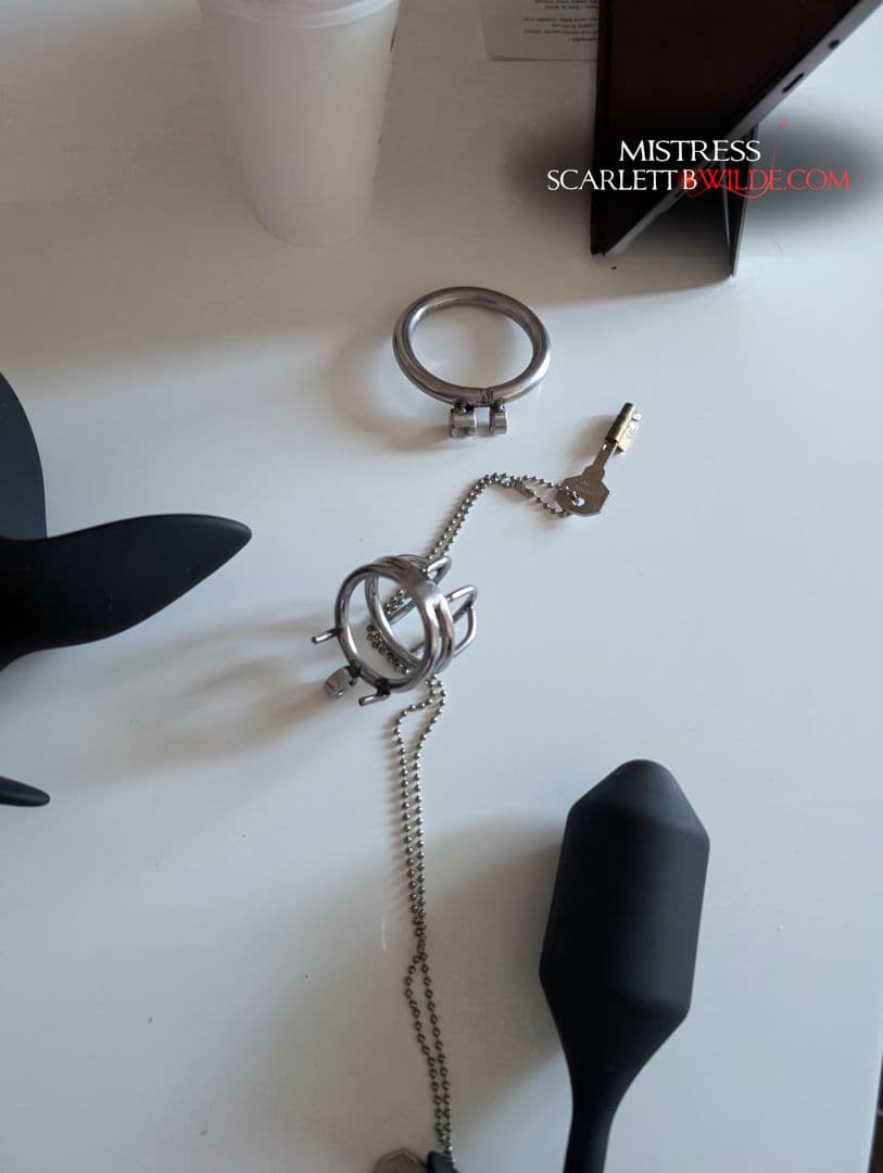 On a desk sits a chastity cock cage, unlocked by key ( on a chain ). Two butt sex toys also rest on table, plus a takeaway coffee cup and laptop.